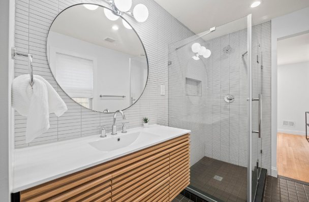 Renovated bathroom with a slatted wood floating vanity, round mirror, white stacked tile, and a glass-enclosed walk-in shower with a niche